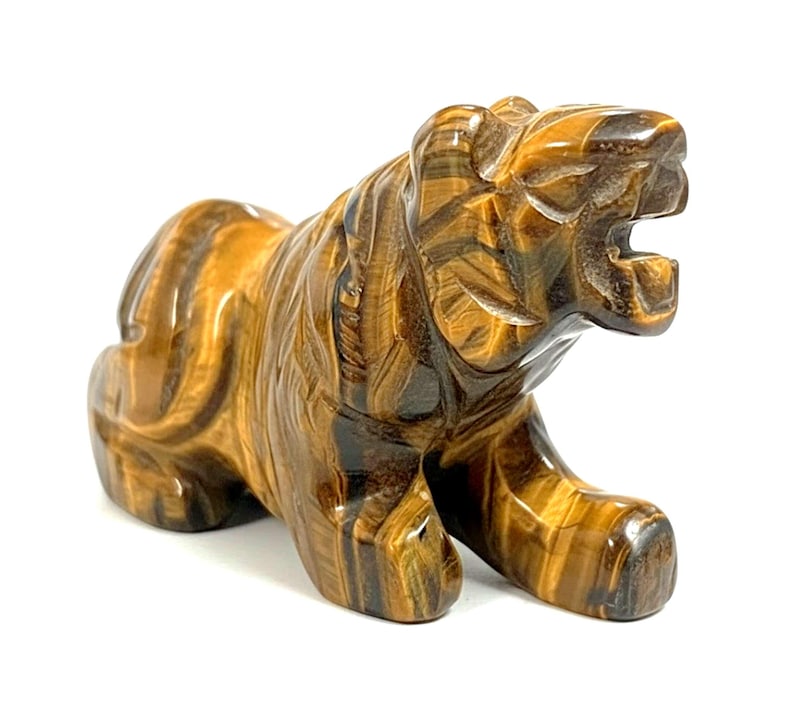 May include: A carved tiger figurine made of tiger's eye stone. The sculpture features a detailed depiction of a tiger in a crouching position with its mouth open. The stone has brown and gold hues.