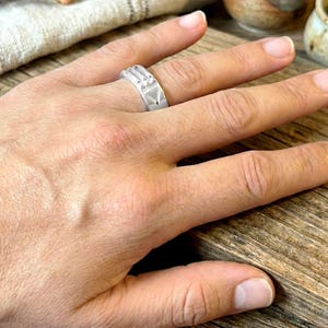 Op de afbeelding: Een zilveren ring met geometrische ontwerpen, waaronder driehoeken en rechthoekige vormen. De ring wordt aan een vinger gedragen, wat de gedetailleerde afwerking en moderne esthetiek benadrukt. De details van de ring zijn zichtbaar.