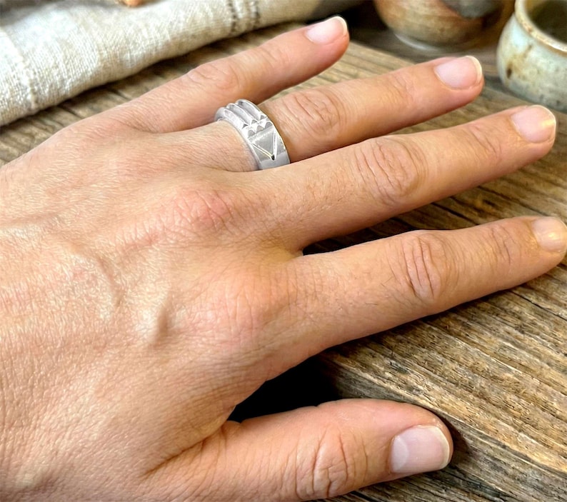 Op de afbeelding: Een zilveren ring met geometrische ontwerpen, waaronder driehoeken en rechthoekige vormen. De ring wordt aan een vinger gedragen, wat de gedetailleerde afwerking en moderne esthetiek benadrukt. De details van de ring zijn zichtbaar.