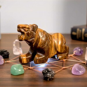 May include: A carved tiger figurine made of tiger's eye stone. The tiger is surrounded by various crystals and stones, including amethyst, quartz, and green aventurine, connected by copper wire. The arrangement is on a wooden surface.