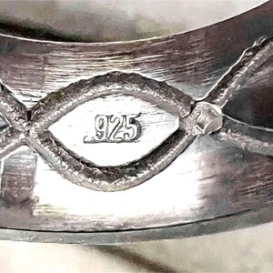 Op de afbeelding: Close-up van een zilveren ring met een getextureerd oppervlak. De ring heeft een decoratief ontwerp met een ruitvormig element in het midden, gemarkeerd met het nummer "925". De ring is gemaakt van sterling zilver.