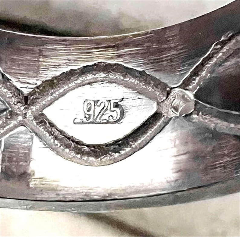 Op de afbeelding: Close-up van een zilveren ring met een getextureerd oppervlak. De ring heeft een decoratief ontwerp met een ruitvormig element in het midden, gemarkeerd met het nummer "925". De ring is gemaakt van sterling zilver.
