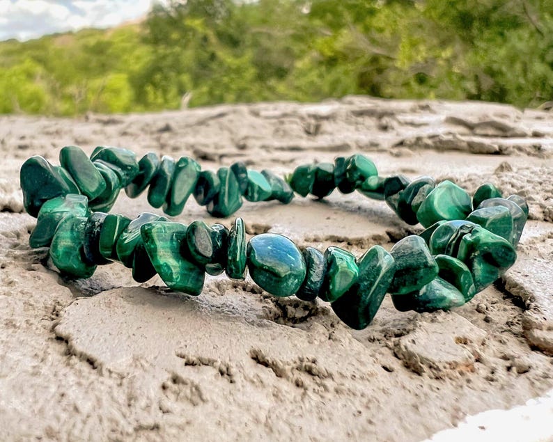 Op de afbeelding: Een armband gemaakt van gepolijste, onregelmatig gevormde malachietstenen. De stenen zijn diepgroen met donkerdere groene adering. De armband ligt op een lichtbruin, getextureerd oppervlak, met een vage groene achtergrond.