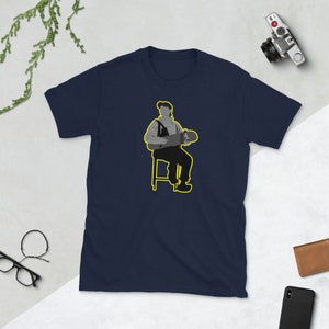 May include: A navy blue t-shirt with a graphic of a person sitting on a stool holding a skateboard. The person is wearing a hat and overalls. The graphic is outlined in yellow.