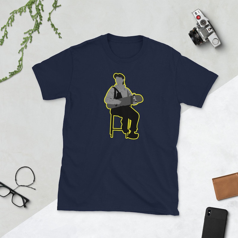 May include: A navy blue t-shirt with a graphic of a person sitting on a stool holding a skateboard. The person is wearing a hat and overalls. The graphic is outlined in yellow.