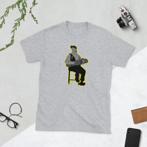 May include: A gray t-shirt with a graphic of a person sitting on a stool holding a skateboard. The person is wearing a black and white outfit and the skateboard is yellow.
