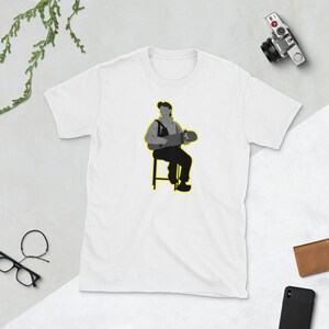 May include: A white t-shirt with a grey silhouette of a person sitting on a stool holding a skateboard. The person is wearing a cap and overalls. The stool and skateboard are outlined in yellow.