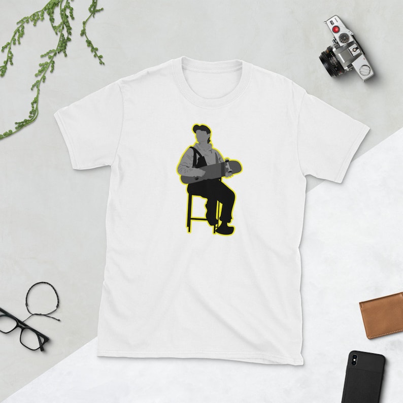 May include: A white t-shirt with a grey silhouette of a person sitting on a stool holding a skateboard. The person is wearing a cap and overalls. The stool and skateboard are outlined in yellow.
