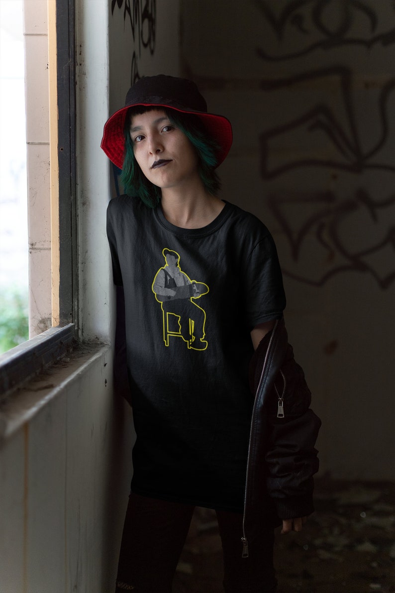 May include: A person wearing a black t-shirt with a yellow outline of a person sitting on a chair holding a paintbrush. The person is wearing a red and black bucket hat and has green hair.
