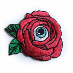 May include: Embroidered patch featuring a red rose with a blue and white eyeball in the center. The rose has green leaves with black outlines. The patch is on a white background.
