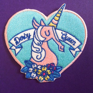 May include: Pink unicorn with a yellow horn and blue mane, embroidered on a light blue heart-shaped patch. The unicorn is surrounded by blue and yellow flowers. The patch has the text "Derby Queen" on a white banner.