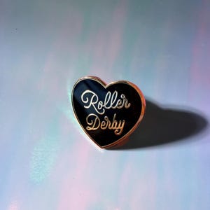 May include: A heart-shaped pin with a black enamel center and gold trim. The pin features the words "Roller Derby" in a cursive font. The pin is set against a light blue and purple background.