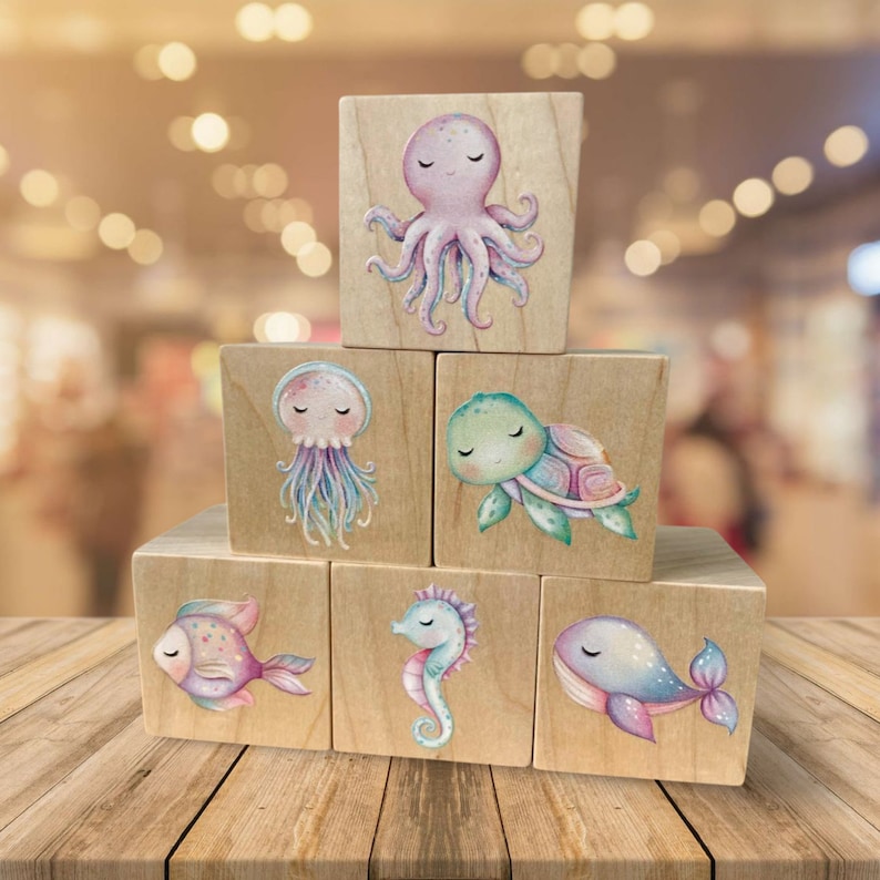 May include: A set of six wooden blocks featuring colorful illustrations of sea creatures. The blocks feature a pink octopus, a blue jellyfish, a green turtle, a purple fish, a blue seahorse, and a blue whale.