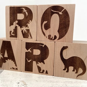 以下が含まれることがあります： 恐竜とヤシの木のシルエットが焼かれた木製のブロックで「ROAR」という単語を綴っています。