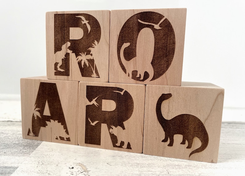 以下が含まれることがあります： 恐竜とヤシの木のシルエットが焼かれた木製のブロックで「ROAR」という単語を綴っています。