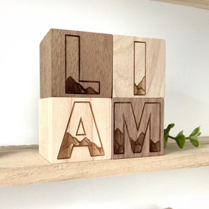 May include: Wooden blocks spelling out the name "Liam" with a mountain range design engraved on each letter. The blocks are arranged in a row on a wooden shelf.