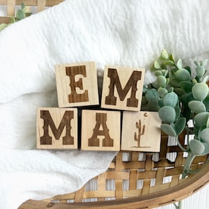 以下が含まれることがあります： 「EMMA」と書かれた木製ブロック。文字はウエスタン風のフォントで彫られています。1つのブロックにはサボテンと三日月のデザインがあります。ブロックは、緑の植物が添えられた白い布の上に配置されています。