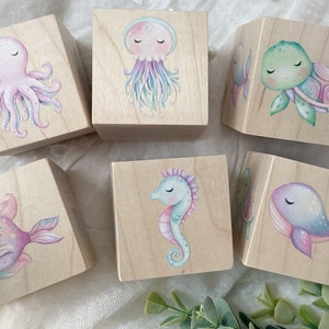 May include: Six wooden blocks with painted sea creatures. Each block features a different creature: an octopus, a jellyfish, a turtle, a fish, a seahorse, and a whale. The blocks are painted in pastel colors and have a whimsical, childlike style.
