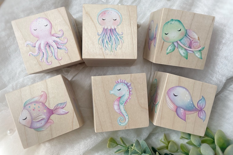 May include: Six wooden blocks with painted sea creatures. Each block features a different creature: an octopus, a jellyfish, a turtle, a fish, a seahorse, and a whale. The blocks are painted in pastel colors and have a whimsical, childlike style.