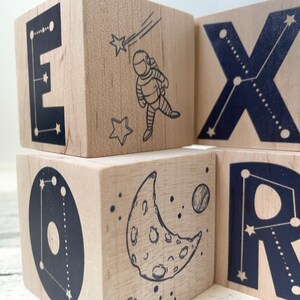 May include: Wooden blocks with black letters spelling out the word "EXPLORER" with a constellation design. Each block features a different space-themed image, including an astronaut, a crescent moon, and a planet.
