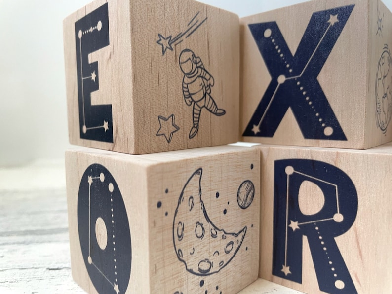May include: Wooden blocks with black letters spelling out the word "EXPLORER" with a constellation design. Each block features a different space-themed image, including an astronaut, a crescent moon, and a planet.
