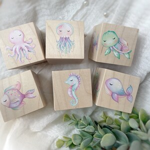 May include: Six wooden blocks with painted sea creatures. The blocks feature a pink octopus, a purple jellyfish, a green turtle, a blue whale, a purple seahorse, and a pink and purple fish.