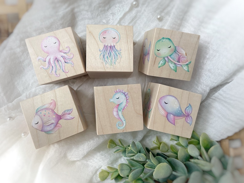May include: Six wooden blocks with painted sea creatures. The blocks feature a pink octopus, a purple jellyfish, a green turtle, a blue whale, a purple seahorse, and a pink and purple fish.