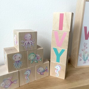 May include: Wooden blocks spelling out the name "IVY" with pink and teal letters. Each block features a different sea creature illustration, including an octopus, a turtle, a jellyfish, a seahorse, and a fish.