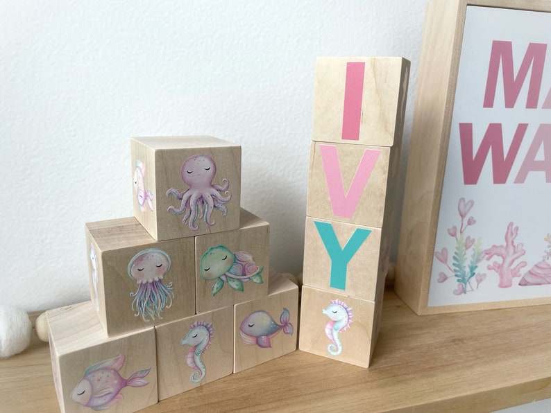 May include: Wooden blocks spelling out the name "IVY" with pink and teal letters. Each block features a different sea creature illustration, including an octopus, a turtle, a jellyfish, a seahorse, and a fish.