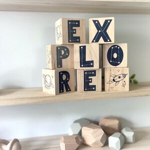 May include: Wooden blocks spelling out the word "EXPLORE" with a constellation design and space-themed illustrations. The blocks are stacked on a wooden shelf.
