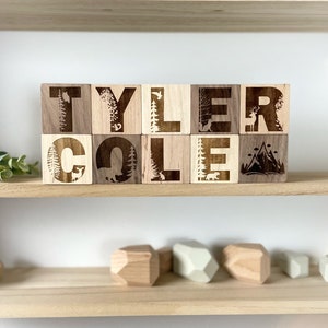 May include: Wooden blocks spelling out the name "TYLER COLE" with a woodland theme. Each block features a different nature-inspired design, including trees, deer, and mountains.