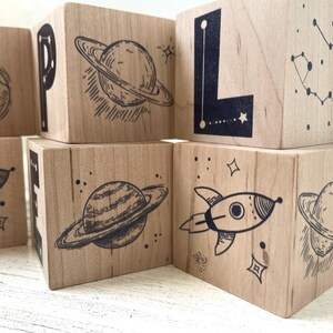 May include: Wooden blocks with a space theme. Each block features a different space-related image, including a planet, a rocket, and a constellation. The blocks are painted in natural wood tones and have black ink illustrations.