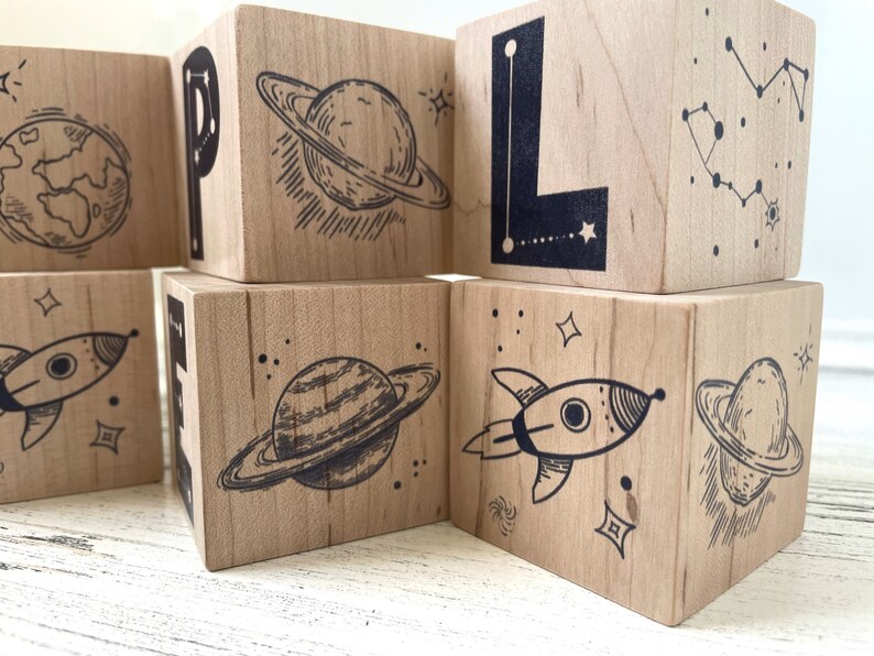 May include: Wooden blocks with a space theme. Each block features a different space-related image, including a planet, a rocket, and a constellation. The blocks are painted in natural wood tones and have black ink illustrations.