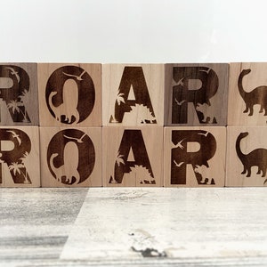 以下が含まれることがあります： 「ROAR」という単語が、大文字で書かれた木製のブロック。各ブロックには、異なる恐竜のシルエットとヤシの木が描かれています。