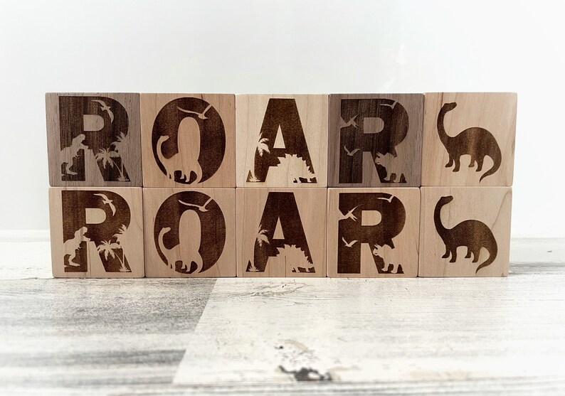 以下が含まれることがあります： 「ROAR」という単語が、大文字で書かれた木製のブロック。各ブロックには、異なる恐竜のシルエットとヤシの木が描かれています。