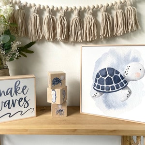 May include: Wooden blocks with a blue and white sea turtle, seahorse, and octopus design stacked on a wooden shelf. A wooden sign with the text "make waves" is on the shelf. A framed watercolor painting of a blue and white sea turtle is on the shelf.