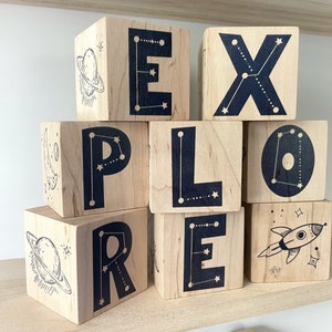 May include: Wooden blocks spelling out the word "EXPLORE" with a constellation design on each block. The blocks are stacked in a pyramid shape. The top block has a rocket ship design.