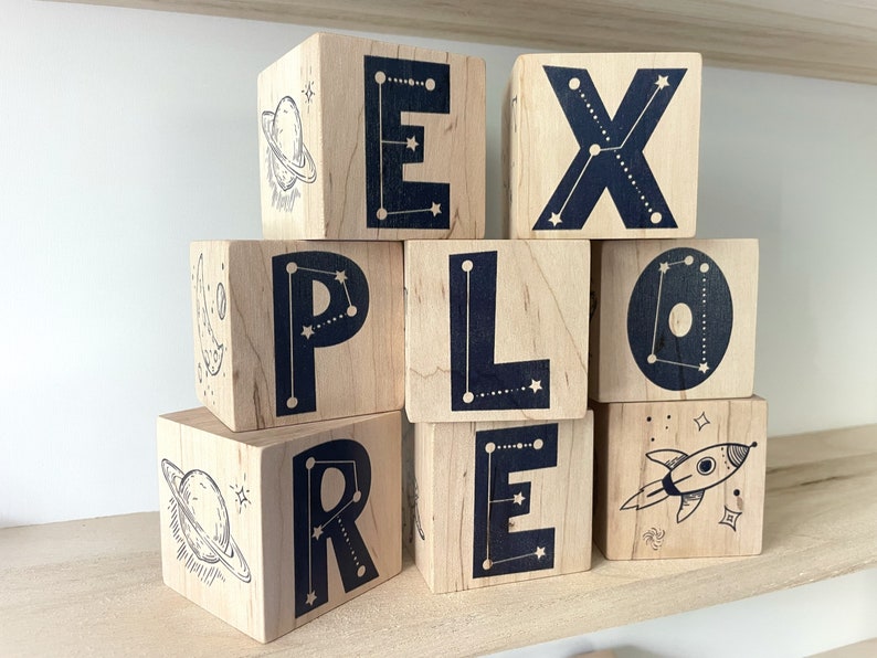 May include: Wooden blocks spelling out the word "EXPLORE" with a constellation design on each block. The blocks are stacked in a pyramid shape. The top block has a rocket ship design.