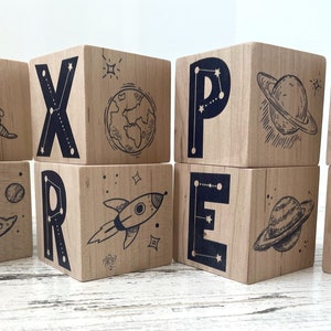 May include: Wooden blocks spelling out the word "EXPLORE" with space-themed illustrations on each block. The illustrations include an astronaut, a rocket, a planet, a moon, and stars.