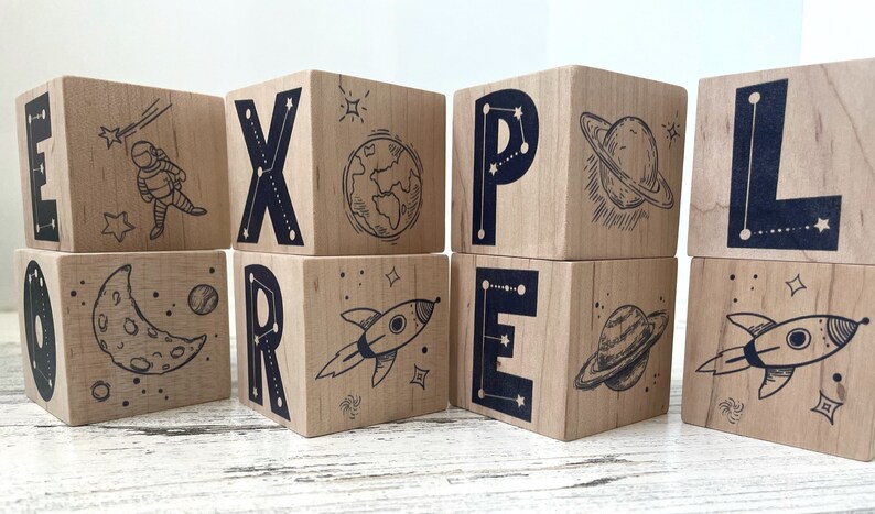 May include: Wooden blocks spelling out the word "EXPLORE" with space-themed illustrations on each block. The illustrations include an astronaut, a rocket, a planet, a moon, and stars.