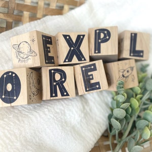 May include: Wooden blocks spelling out the word "EXPLORE" with constellation and space-themed illustrations. Each block features a different letter and a unique design, including a rocket, a crescent moon, a planet, and stars.