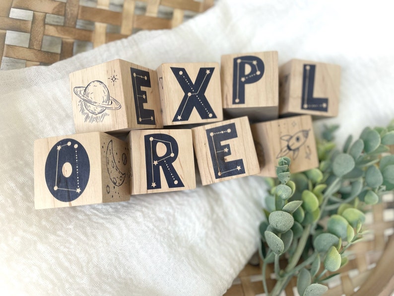 May include: Wooden blocks spelling out the word "EXPLORE" with constellation and space-themed illustrations. Each block features a different letter and a unique design, including a rocket, a crescent moon, a planet, and stars.