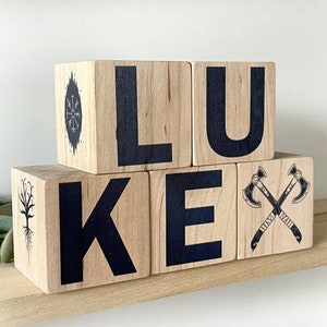 以下が含まれることがあります： 黒い文字と装飾的なイラストで「LUKE」と綴られた木製ブロック。ブロックは薄茶色で、斧、木、羅針盤の黒いイラストが描かれています。ブロックは薄茶色の棚に飾られています。