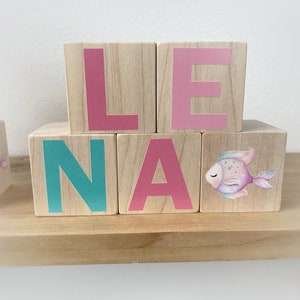 May include: Wooden blocks spelling out the name "Lena" with a pink, teal, and pink colour scheme. The "A" block has a watercolour fish design.
