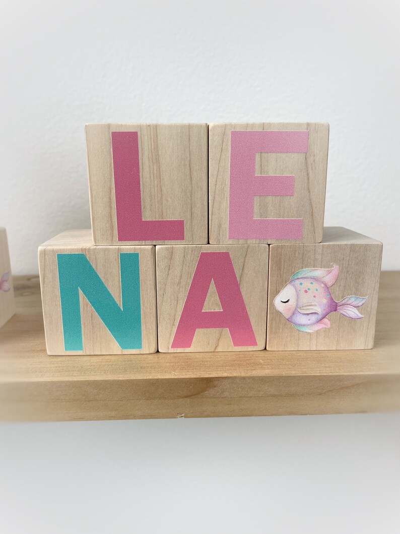 May include: Wooden blocks spelling out the name "Lena" with a pink, teal, and pink colour scheme. The "A" block has a watercolour fish design.