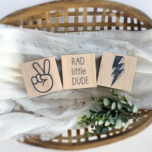 May include: Three wooden blocks with different designs. The first block has a peace sign, the second block says "RAD little DUDE", and the third block has a lightning bolt.