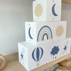 May include: A stack of white wooden blocks with different blue and beige illustrations. The blocks feature a sun, moon, rainbow, cloud, hot air balloon, and shooting star.