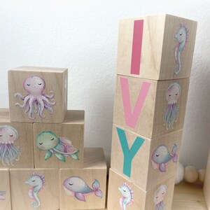 May include: Wooden blocks spelling the name "IVY" with pink, teal, and white letters. Each block features a different sea creature illustration, including an octopus, turtle, jellyfish, whale, and seahorse.