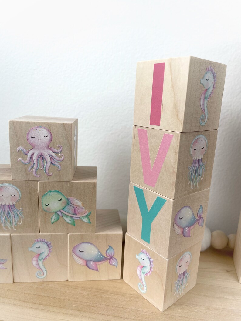 May include: Wooden blocks spelling the name "IVY" with pink, teal, and white letters. Each block features a different sea creature illustration, including an octopus, turtle, jellyfish, whale, and seahorse.