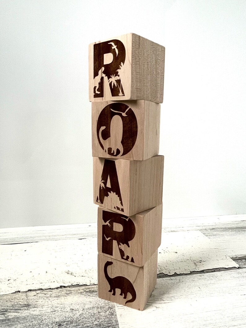 以下が含まれることがあります： 恐竜のシルエットとヤシの木が描かれた木製のブロックで「ROAR」という単語を綴っています。